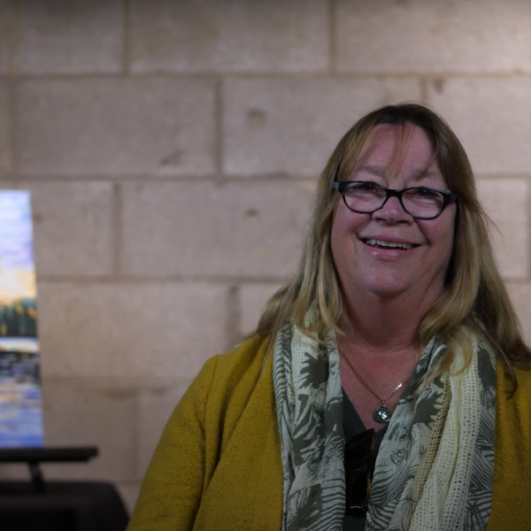 A photo of artist Diane Thoms sitting in front of her painting of an island on the water.