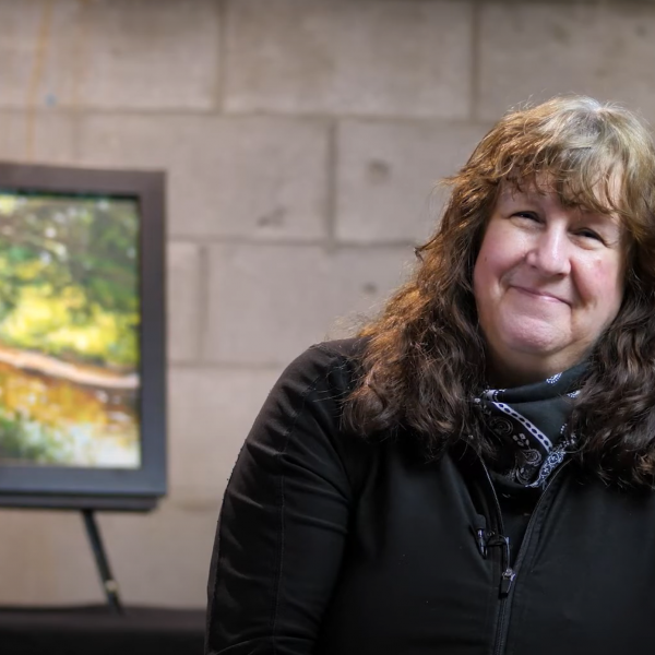 A photo of artist Janine Marson smiling with her painting in the background.